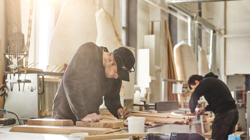 Zwei Handwerker arbeiten in einer Werkstatt an der Veredelung von Holzelementen für ein Sideboard aus Holz, unterstützt durch moderne Outsourcing-Strategien.