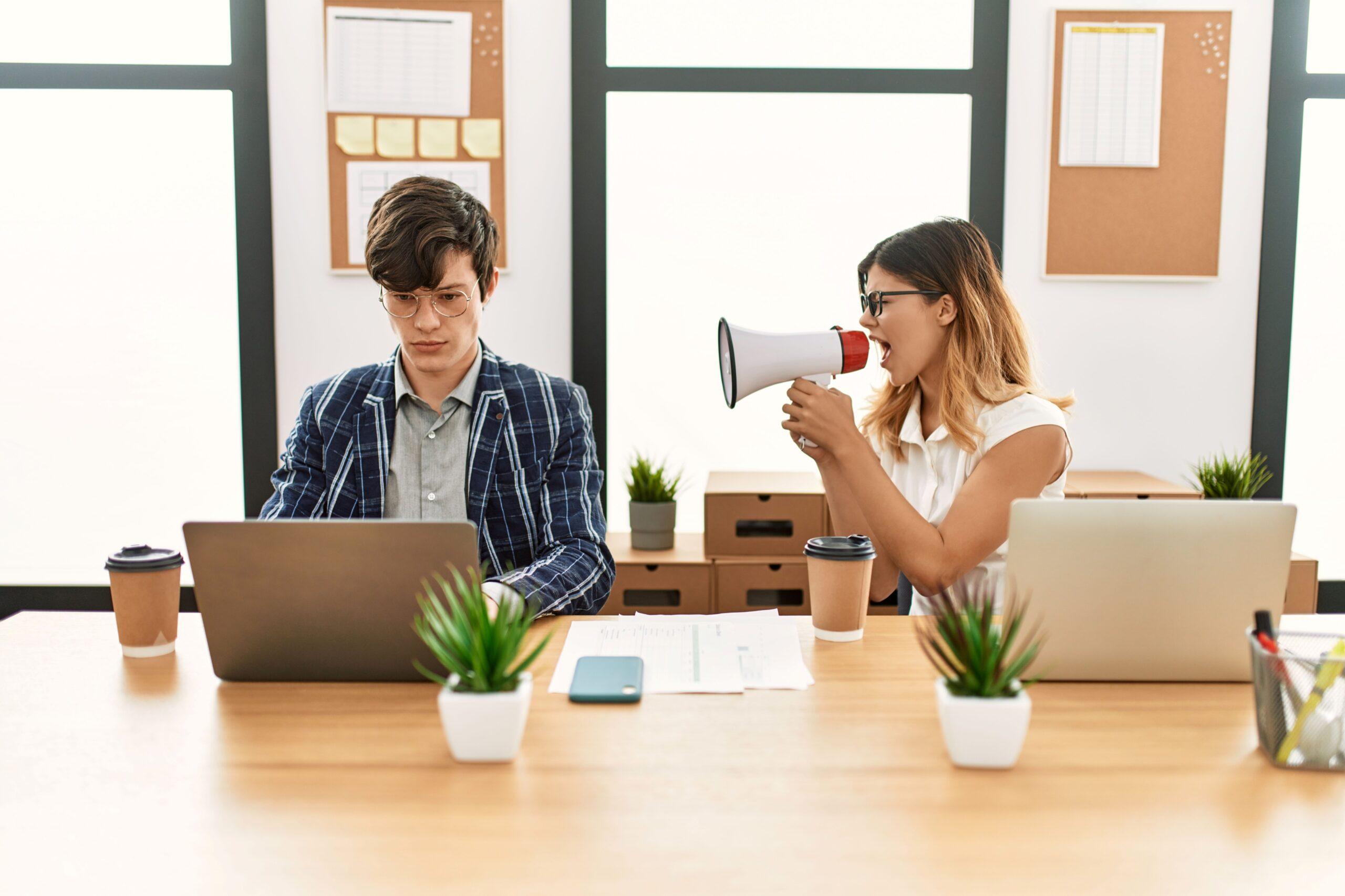 Zwei Personen sitzen an einem Tisch. Eine Peron ist am arbeiten, die andere sitzt mit einem Megafon neben ihr 