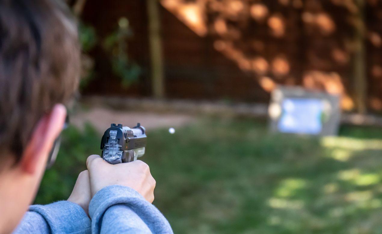 Teilnehmer zielt mit Softair-Waffe bei Outdoor-Teamspiel auf Zielscheibe im Garten, Fokus auf Konzentration und Taktik